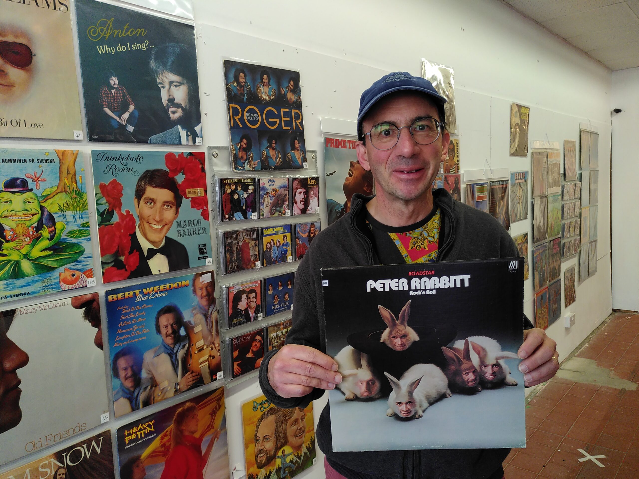 Man stood in a gallery holding an album cover for Roadstar Peter Rabbitt, featuring five rabbits with band members faces superimposed on rabbits' heads.