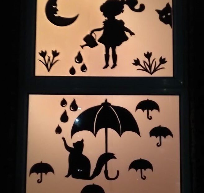 An illuminated window art image of a girl under a moon pouring water from a watering can. A cat is pawing at the drops of water